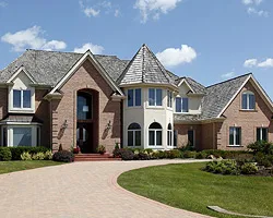 Large brick house with a paved driveway, a round bay with a turret and a lawn with plantings